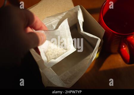 Eine Hand greift aus einer offenen Box neben einem roten Becher auf einem Holztisch im goldenen Stundenlicht nach einem Teebeutel. Schwarzweiß-Bild. Stockfoto