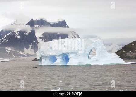 Zwei Crew in einem Tierkreisboot vom Le Boreal-Kreuzschiff passieren einen riesigen Eisberg im Südatlantik, südwestlich der Petermann-Insel, Antarktis. Stockfoto