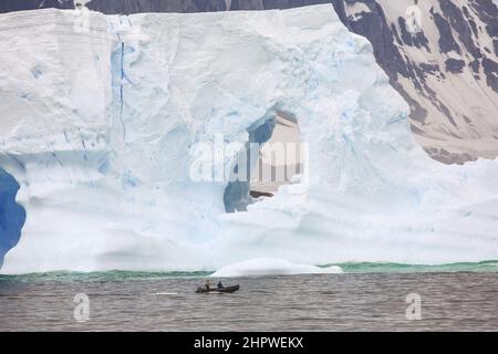 Zwei Crew in einem Tierkreisboot vom Le Boreal-Kreuzschiff passieren einen riesigen Eisberg im Südatlantik, südwestlich der Petermann-Insel, Antarktis. Stockfoto