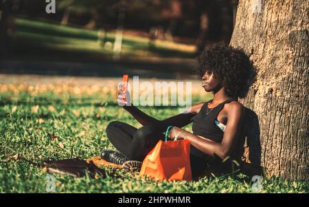 Eine Seitenansicht einer schönen, lockigen afrikanischen Frau, die auf dem Rasen sitzt und sich an einen Parkbaum lehnt, während sie einen Videoanruf mit ihrem Freund V führt Stockfoto