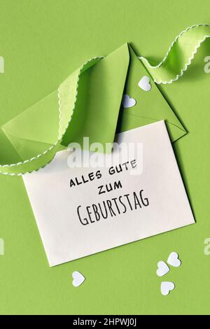 Alles gute zum Geburtstag bedeutet Happy Birthday Text auf der Karte. Frühlingshintergrund in grün. Papierumschlag mit Grußkarte und Herzkonfetti auf grün Stockfoto