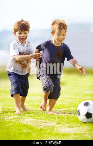 Mit einem freundlichen Spiel. Zwei süße kleine Jungen, die draußen zusammen Fußball spielen, während sie von Schlamm bedeckt sind. Stockfoto