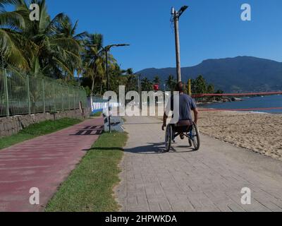 Ein behinderter Mann im Rollstuhl nähert sich dem Betrachter. Er nutzt den Fußgängerweg auf der Insel Ilhabela, Brasilien, was sehr praktisch ist Stockfoto
