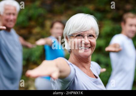 Yoga bringt mich zu meinem glücklichen Platz. Aufnahme einer älteren Frau, die mit anderen Menschen im Freien Yoga macht. Stockfoto