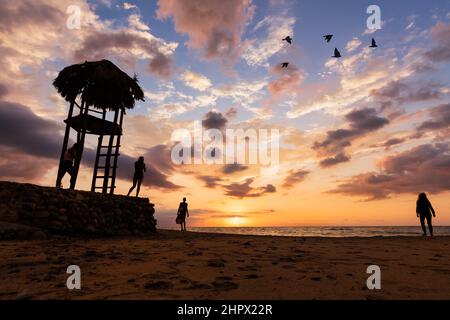 Hochauflösendes Bild eine Ozeanlandschaft mit Silhouetten von Menschen am Strand und Einer Hütte, die auf Einer Steinmauer sitzt Stockfoto