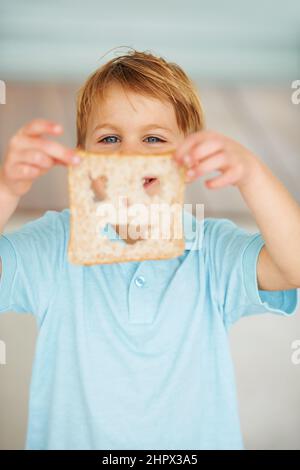 Das Essen macht mich glücklich. Aufnahme eines kleinen Jungen, der eine Scheibe Brot mit einem ausgeschnittenen Smiley-Gesicht hochhält. Stockfoto