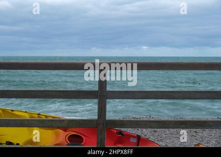 Farbenfrohes Kajak auf einem Kiesstrand, das durch das Geländer am blaugrünen Ozean und dem stürmischen Himmel gesehen wird Stockfoto