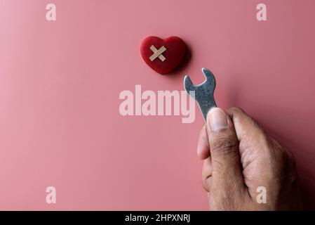 Reparieren, reparieren ein gebrochenes Herzkonzept. Rotes Herz, Verband und Schraubenschlüssel auf rosa Hintergrund mit Kopierfläche. Stockfoto