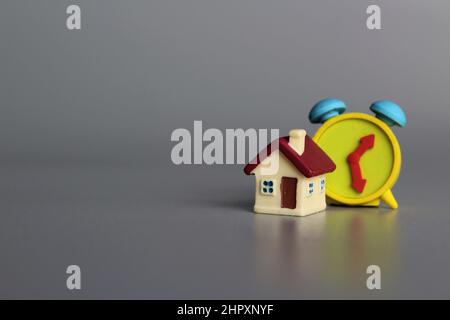 Spielzeughaus und Wecker auf grauem Hintergrund mit Platz für Text. Immobilienkonzept. Stockfoto