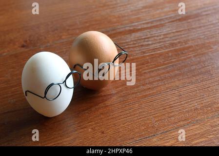Ähnliches, aber anderes Konzept. Weißes Ei und braunes Ei tragen Gläser auf Holztisch Stockfoto