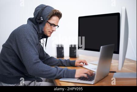 In seine Arbeit vertieft. Ein junger Hacker, der auf zwei Bildschirmen und einem Laptop mit Hoodie arbeitet. Stockfoto