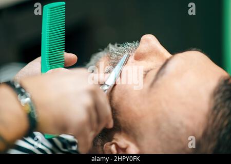 Männlicher Kunde in einem Friseurladen, der einen Bart- und Schnurrbart-Besatz aus der Nähe auf dem Kamm und eine Schere in den Händen des Barbers hat Stockfoto