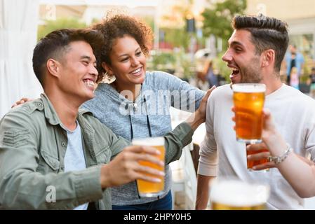 Gruppe von fröhlichen multirassischen Freunden, die einander beim Trinken von alkoholischem Bier auf der Terrasse des Cafés im Freien in der Stadt betrachten Stockfoto