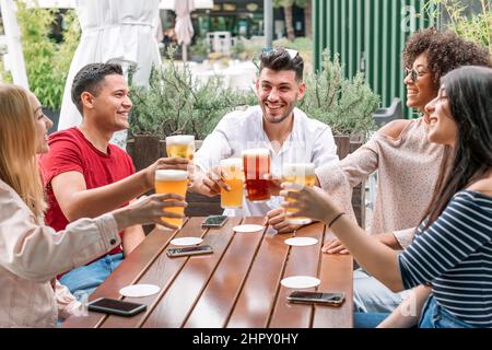 Gesellschaft von fröhlichen jungen multirassischen Freunden, die mit Gläsern Bier toasten, während sie die Wiedervereinigung auf der Terrasse des Restaurants am Sommertag feiern Stockfoto