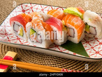 Appetitliche Uramaki-Sushi-Rollen in Regenbogenfarben mit Krabben und Avocado, gekrönt von verschiedenen Fischsorten, die in der Nähe von Essstäbchen auf dem Teller serviert werden Stockfoto