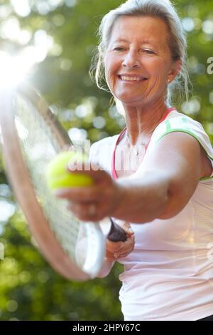 Ich hatte jahrelange Praxis, um meinen Dienst zu perfektionieren. Lächelnde ältere Frau, die sich zum Dienst bereit gemacht hat - Tennis. Stockfoto