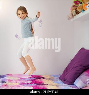 Hüpfend vor Freude. Porträt eines niedlichen kleinen Mädchens, das zu Hause auf ihrem Bett springt. Stockfoto