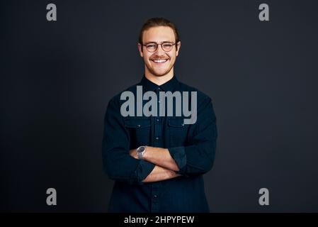 Seien Sie darauf vorbereitet, sich im Geschäft abzuheben. Ein Porträt eines hübschen jungen Geschäftsmannes, der allein steht und die Arme vor einem schwarzen Hintergrund gefaltet hat Stockfoto