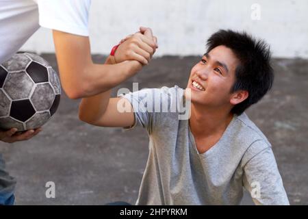 Eine helfende Hand geben. Asiatischer Junge, der seinem Freund während eines Fußballspiels vom Boden aus hilft. Stockfoto