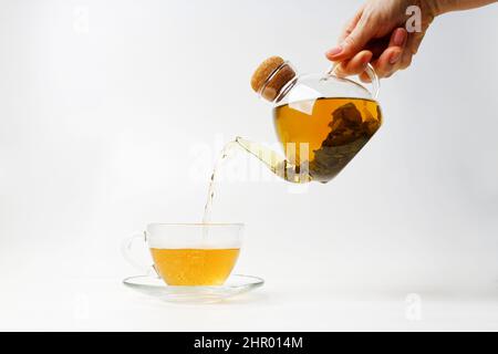 Menschliche Hand gießt Tee aus einem durchsichtigen Wasserkocher in eine transparente Tasse auf weißem Hintergrund. Stockfoto