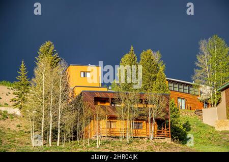 Moderne Holzhäuser mit Blick auf den Hang, umgeben von Espenbäumen unter dunkelblauem ominösen Himmel im Sommer. Stockfoto
