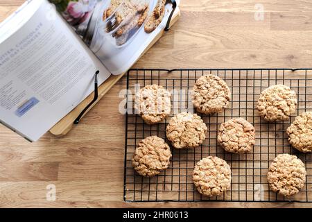 Eine Charge frisch gebackener Plätzchen, die auf einem Kühlregal auf einer Küchenarbeitsfläche kühlen. Ein Buch mit dem Rezept liegt auf einer Seite der Kekse Stockfoto