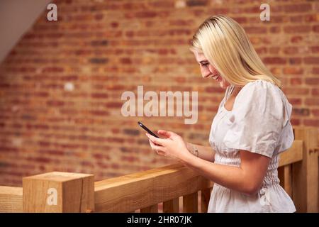 Frau, Die Zu Hause Am Holztreppe Mit Dem Mobiltelefon Steht Stockfoto