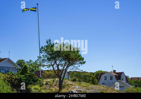 Die schwedische Nationalflagge fliegt auf einem Fahnenmast im Wind auf einem Hügel in einer Siedlung von Landhäusern auf der Südkoster-Insel, Bohuslän, Schweden. Stockfoto