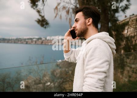 Trauriger junger Mann, der am Telefon spricht, junger Mann mit weißem Sweatshirt führt ein stressiges Gespräch, bärtiger junger Mann steht gegen die Ansicht Stockfoto