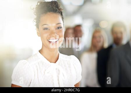 Erfolg bringt mir immer ein Lächeln ins Gesicht. Zugeschnittenes Porträt einer jungen Geschäftsfrau, die in ihrem Büro mit Kollegen im Hintergrund steht. Stockfoto