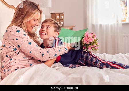 Liebevoller Sohn Überraschende Mutter im Bett mit Blumen und hausgemachter Karte zum Geburtstag oder Muttertag Stockfoto