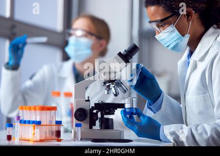Zwei Laborarbeiterinnen, die eine persönliche Schutzausrüstung tragen und im Labor mit Mikroskop an der Erforschung des Covid-19-Impfstoffs arbeiten Stockfoto