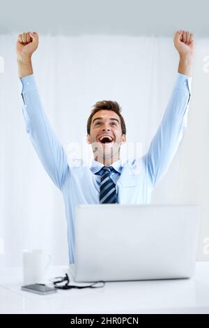 Er hätte nie gedacht, dass er die Frist einhalten muss... Aber er tat es. Ein Geschäftsmann, der mit seinem Computer vor ihm an seinem Schreibtisch sitzt und mit seinen Armen jubelt Stockfoto
