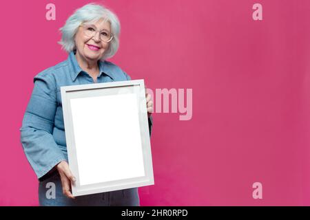 Grauhaarige ältere Frau mit leerem Bilderrahmen auf rosa Hintergrund Stockfoto