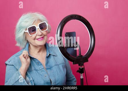 Porträt einer älteren Frau mit Retro-Sonnenbrillen, die in Denim-Overalls vor dem Mobiltelefon posiert, und einem hellen Ring auf rosa Hintergrund Stockfoto