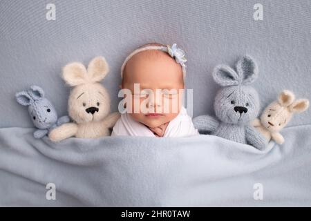 Schlafendes neugeborenes Mädchen in einem weißen Kokon mit weißem Verband und einer blauen Blume. Studio Makrofotografie, Porträt einer neugeborenen Ballerina, Spielzeug Hasen. Stockfoto