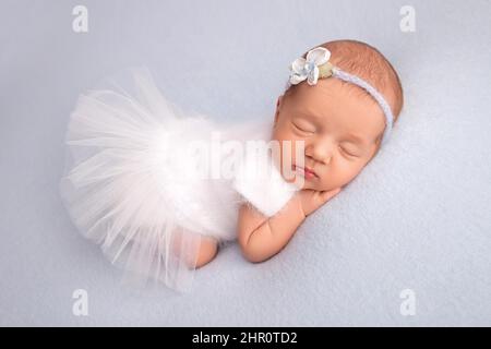 Schlafendes neugeborenes Mädchen in einem weißen Ballettkleid mit einem weißen Verband und einer blauen Blume.Porträt einer neugeborenen Ballerina. Stockfoto