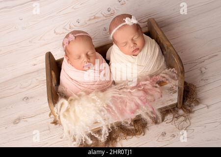 Kleine neugeborene Zwillingsmädchen in Holzkrippe vor dem Hintergrund alter weißer Vintage-Bretter. Neugeborene Zwillingsmädchen mit weißen und rosa Stirnbändern mit Schleifen Stockfoto