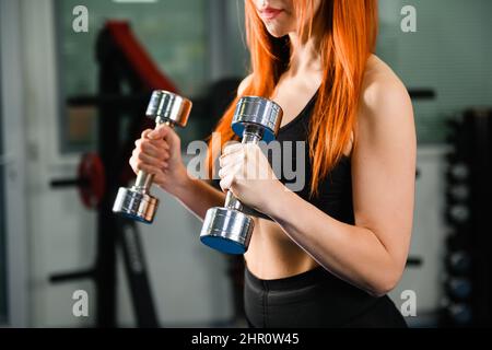 Weiblicher sportlicher Körper in Sportbekleidung mit Hantelgewichten Dumphanteln im modernen dunklen Gym-Interieur, gesunder Lebensstil Stockfoto