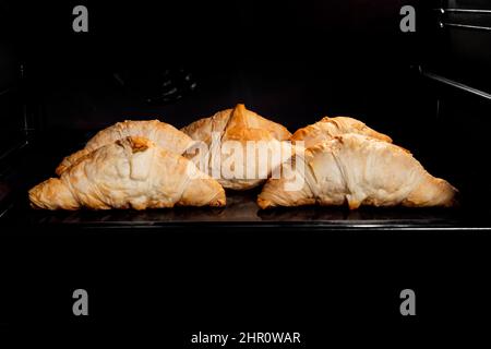 Sechs gebackene hausgemachte Croissants auf Tablett im elektrischen Ofen, schwarzer Hintergrund Stockfoto