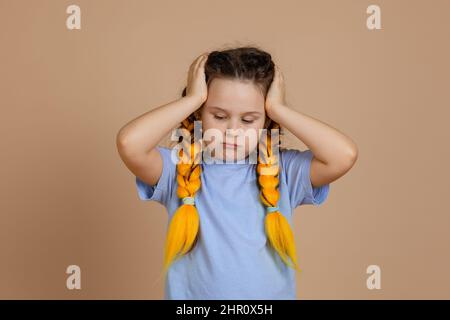 Verärgert kaukasischen kleinen weiblichen Kind hält den Kopf mit den Händen nach unten mit Kanekalon Zöpfe der gelben Farbe auf dem Kopf in blauem T-Shirt auf beige schauen Stockfoto