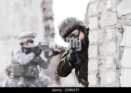 Dynamische Verteidigungskraft. Ein Soldat gucken um eine Wand, während er während einer Rade auf ein beschädigtes Gebäude mit einem Gewehr auf die Kamera zeigt. Stockfoto