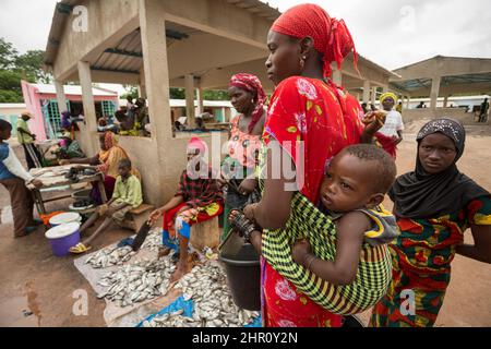 Eine Mutter trägt ihr Baby auf dem Rücken, während sie auf einem Markt in Tanaff, Senegal, Westafrika, frischen Fisch kauft. Stockfoto