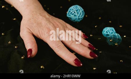 Reife Frauenhand mit rot polierten Nägeln auf schwarzem Hintergrund mit Dekor. Nicht erkennbare Person mit Maniküre beschneiden. Konzept von Pflege und Schönheit Stockfoto