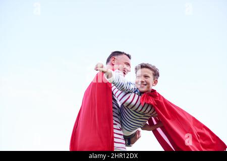 In das wilde Blau des Jenseits. Aufnahme eines Vaters und seines jungen Sohnes, der sich beim Spielen im Freien als Superhelden ausgibt. Stockfoto