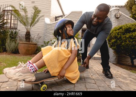 Glücklicher afroamerikanischer Vater schiebt Tochter auf Skateboard im Hinterhof während des Wochenendes Stockfoto