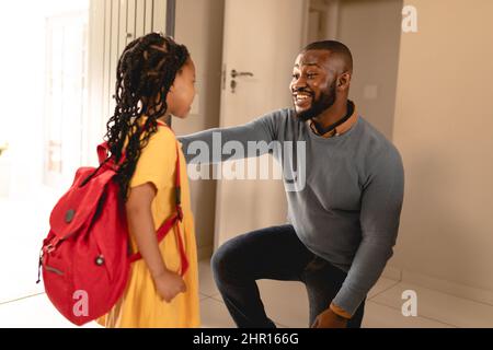Ein glücklicher afroamerikanischer Mann kniete, während er mit seiner Tochter sprach, die am Hauseingang zur Schule ging Stockfoto