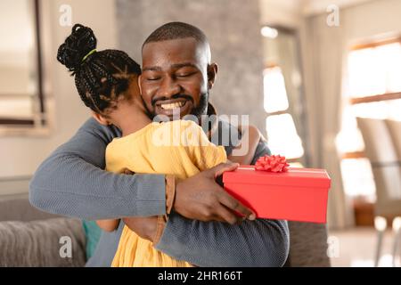 Glücklicher afroamerikanischer Vater umarmte Tochter, nachdem er von ihr zu Hause rotes Geschenk erhalten hatte Stockfoto