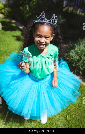 Hochwinkelporträt eines lächelnden Biracial-Mädchens, das eine Tiara-Krone trägt und einen Sternstab im Hinterhof hält Stockfoto