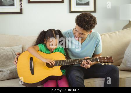 Glücklicher Biracial Vater lehrt Tochter Gitarre spielen, während zusammen auf dem Sofa im Wohnzimmer sitzen Stockfoto